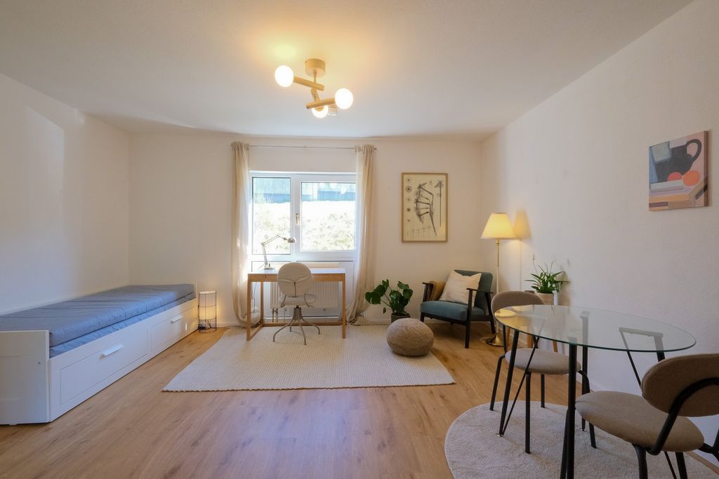Helles, modernes Wohnzimmer mit einem Sofa, einem Schreibtisch vor einem Fenster, einem Stuhl, einem runden Glas-Tisch mit Stühlen und einer Stehlampe.