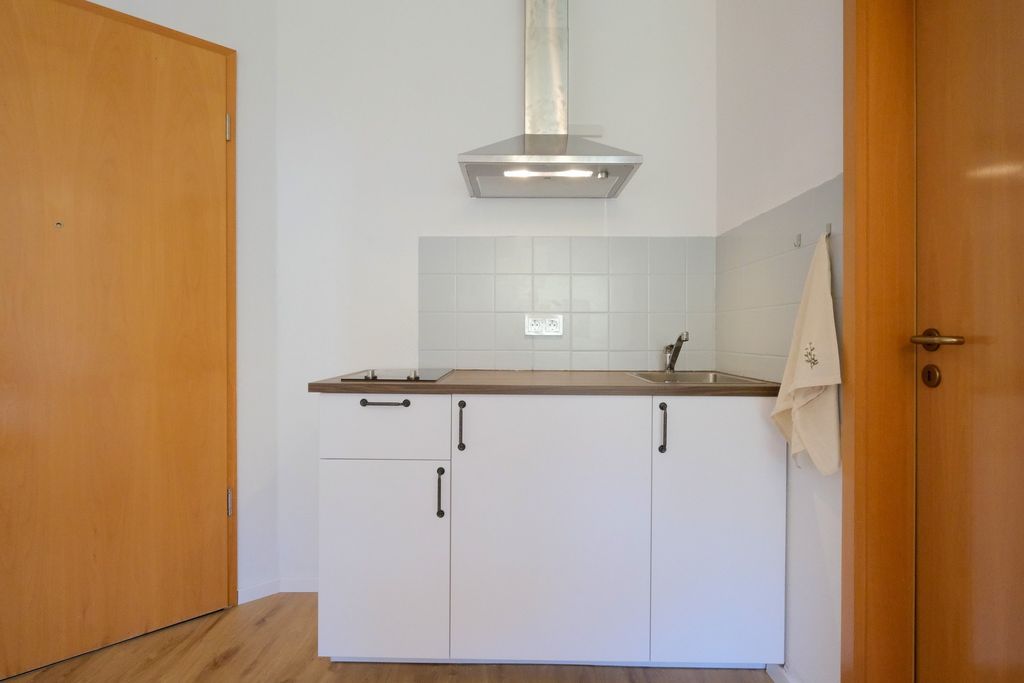 Kleine moderne Küchenzeile mit weißer Front, brauner Arbeitsfläche, Edelstahl-Dunstabzugshaube, Einbaukochfeld und Spüle, zwei Holztüren rechts und links.