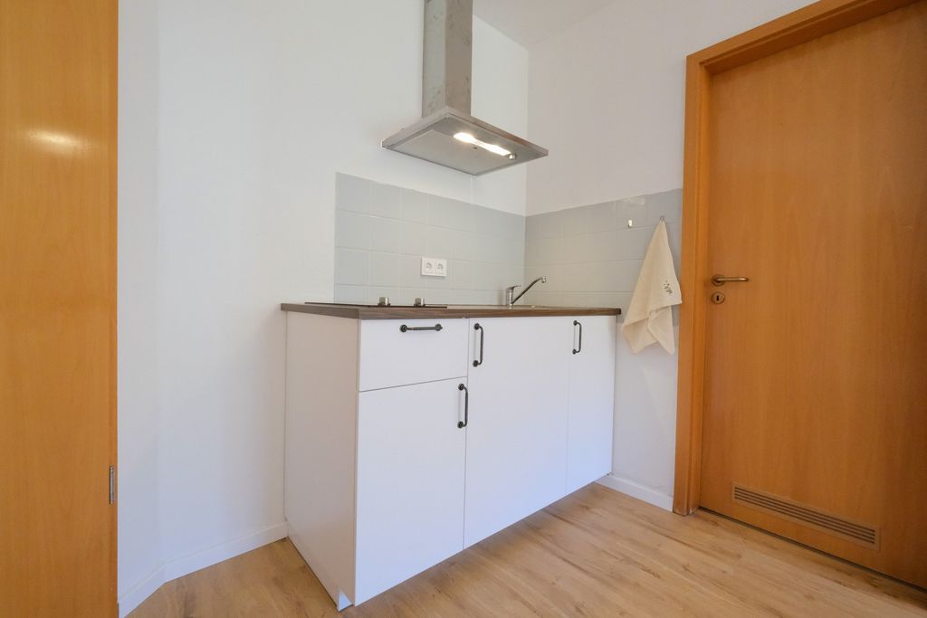 Kleine Küche mit weißem Küchenschrank, Holzboden, Dunstabzugshaube und Holztür in einem schlichten, hellen Raum.