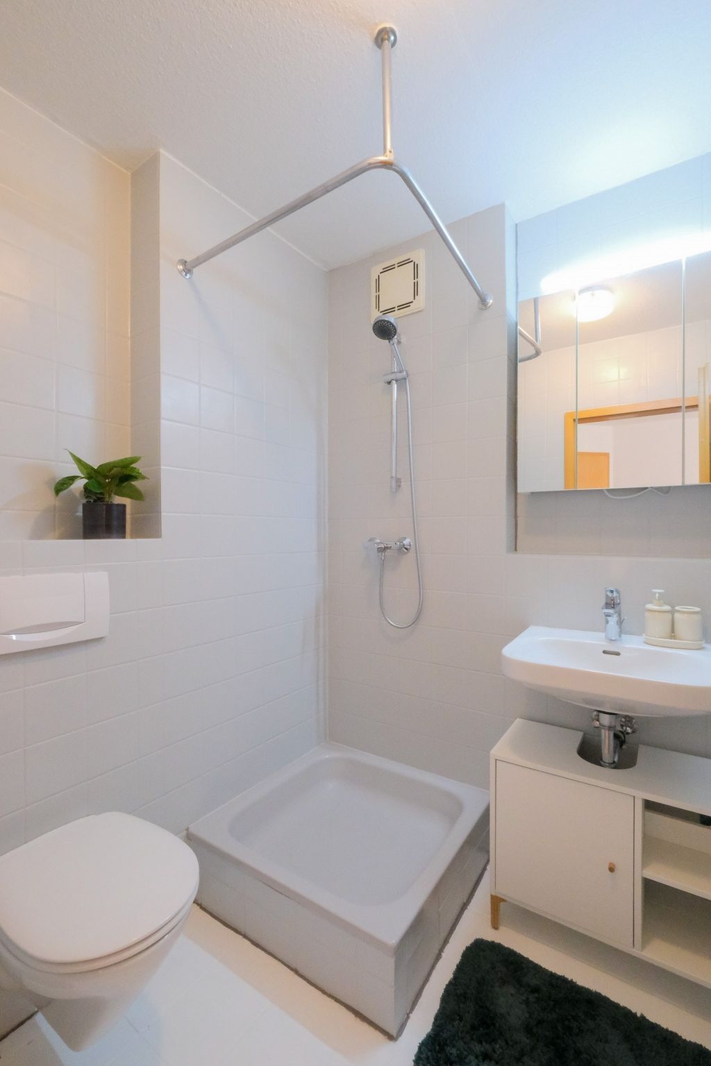 Kleine, minimalistische Badezimmer mit Duschkabine, weiße Wandfliesen, Toilette, Waschbecken mit Unterschrank, Spiegel und einem kleinen Pflanzenkübel auf einer Wandnische.