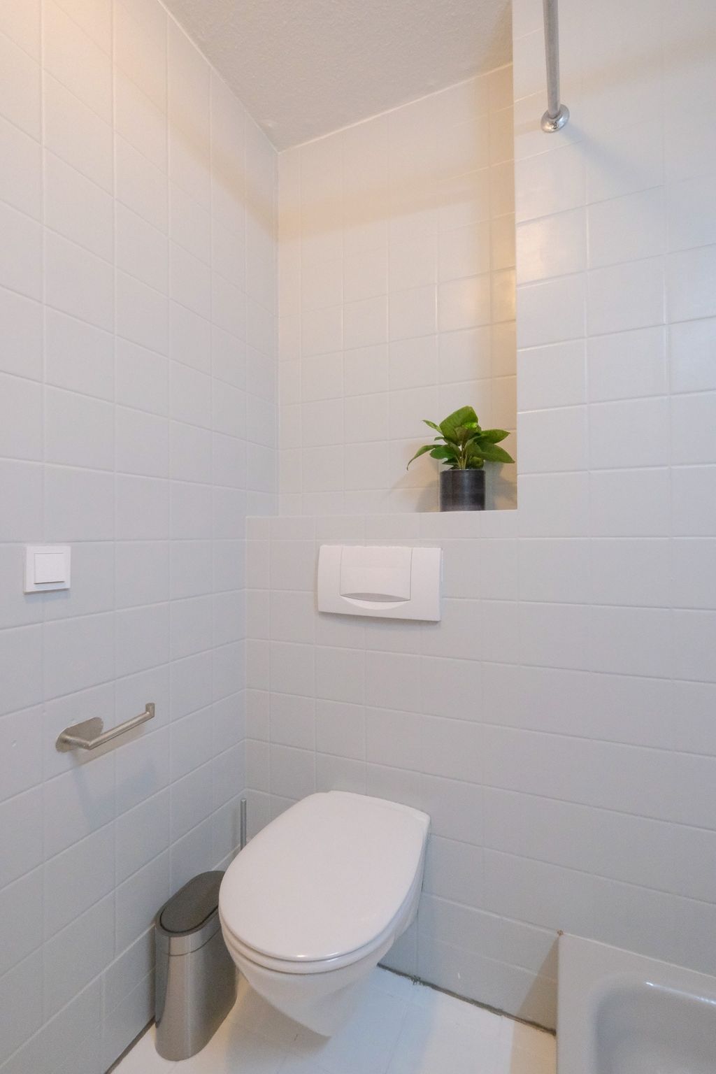 Kleines, modernes Badezimmer mit weiß gekachelten Wänden, wandhängendem WC, Metall-Mülleimer und einem kleinen grünen Topfpflanze in einer Wandnische.