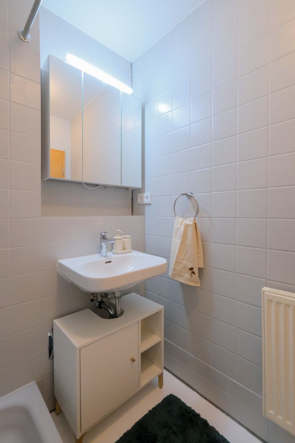 Kleines Badezimmer mit weißem Waschbecken auf einem weißen Unterschrank, Wandspiegel mit Leuchte und einem Handtuch an einem Handtuchring an der Wand.