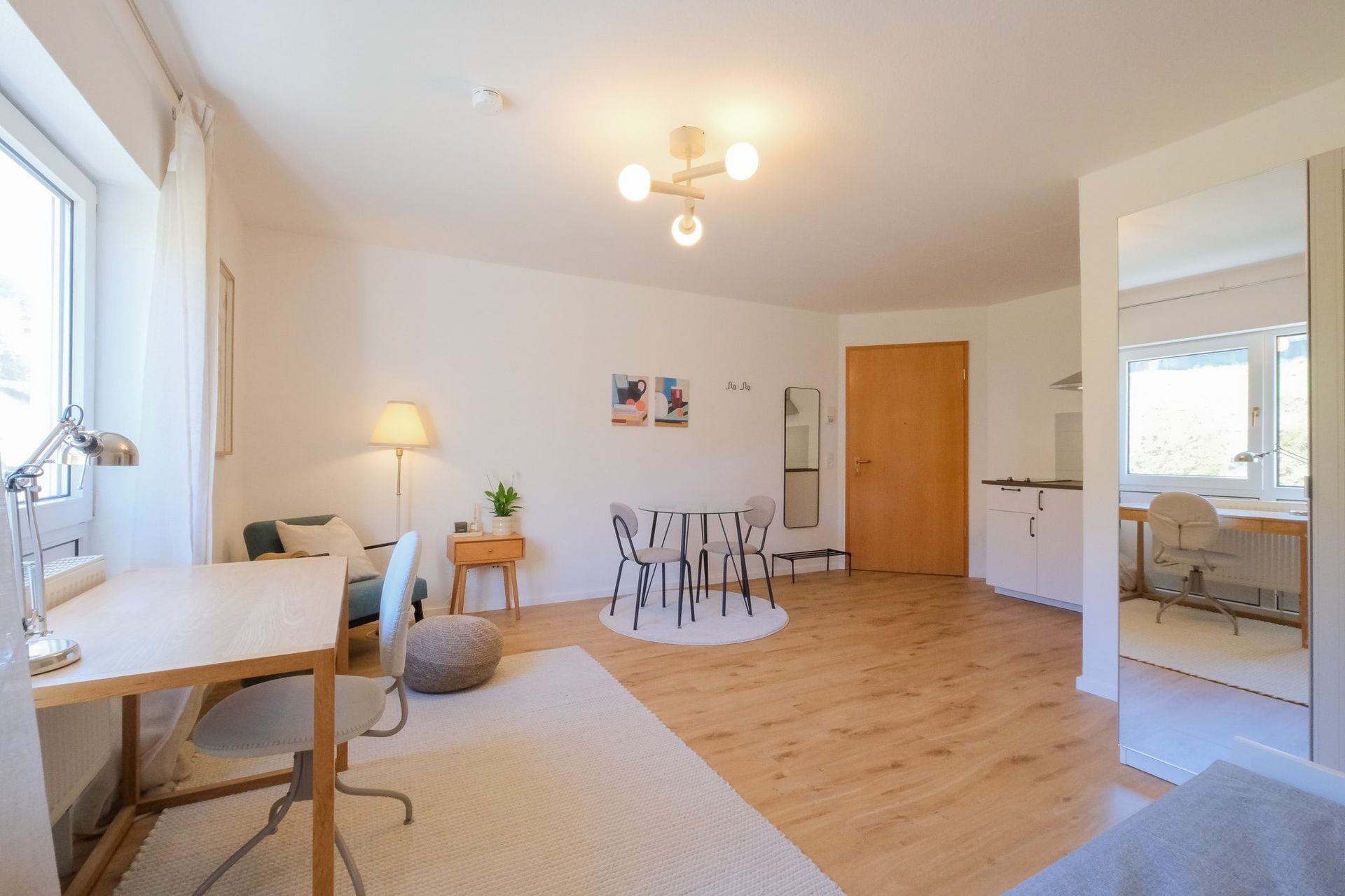 Hell eingerichtetes Wohnzimmer mit Holzfußboden, einem runden Esstisch mit vier Stühlen und einem Arbeitsbereich mit Schreibtisch und Stuhl am Fenster.