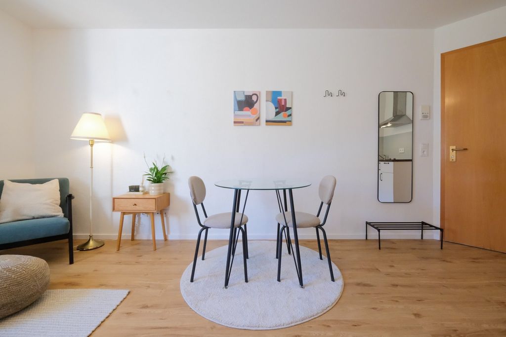 Ein heller Essbereich mit rundem Glastisch und zwei Stühlen auf einem runden beigefarbenen Teppich vor einer weißen Wand mit zwei abstrakten Bildern. Links steht eine Stehlampe neben einem kleinen Holztisch mit Pflanze, rechts ist eine Holzhaustür und eine Wandspiegel mit Ablage.