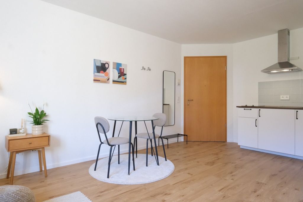 Ein heller Wohnraum mit einem kleinen runden Essbereich in der Mitte, bestehend aus einem weißen runden Tisch und drei grauen Stühlen auf einem runden Teppich. Im Hintergrund befindet sich eine Holzhaustür, ein Wandspiegel und eine minimalistische Küchenzeile mit einem Dunstabzug.
