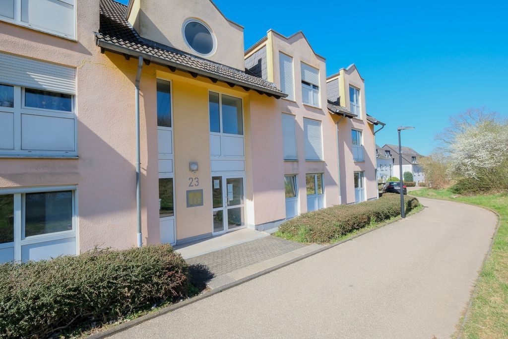 Mehrfamilienhaus mit rosa Fassade und weißen Fenstern an einem sonnigen Tag neben einem mit Sträuchern gesäumten Gehweg.