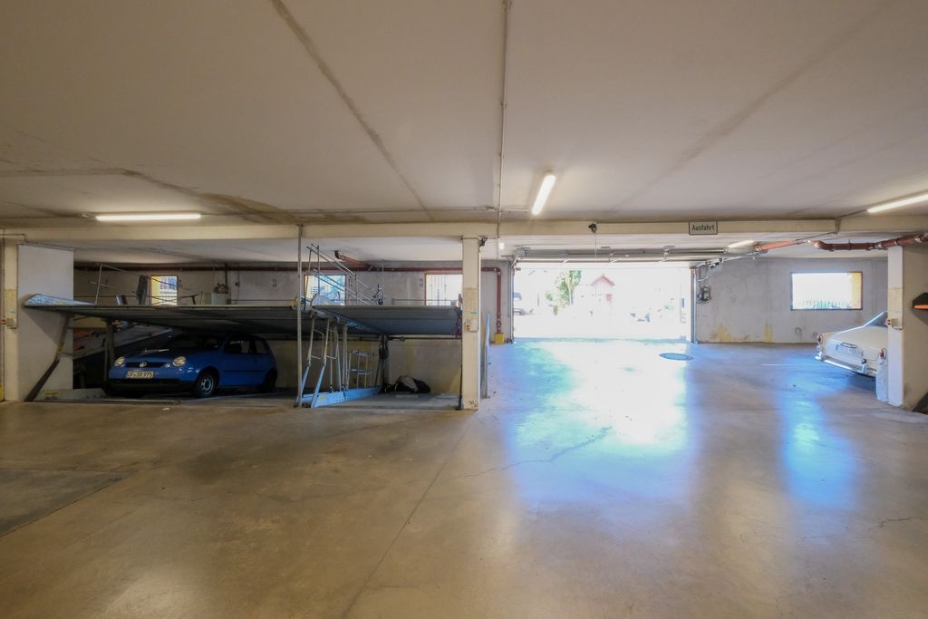 Ein großer, leerer Parkbereich in einer Garage mit zwei geparkten Autos, eines blau links und eines weiß rechts, und einer halb geöffneten Garage im Hintergrund.