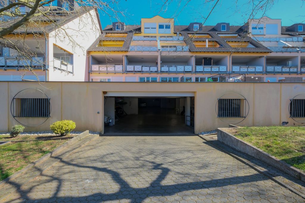 Einfahrt zu einer Tiefgarage vor einem mehrstöckigen Wohngebäude bei sonnigem Wetter mit sichtbaren Baumästen, die Schatten auf den Boden werfen.
