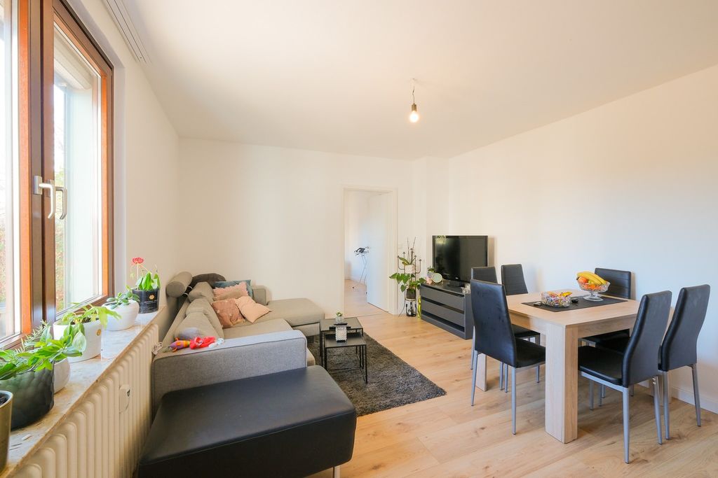 Ein helles Wohnzimmer mit einem grauen Ecksofa, einem schwarzen Hocker, einem Esstisch mit sechs schwarzen Stühlen und einem Flachbildfernseher auf einem Sideboard, beleuchtet durch ein großes Fenster links.