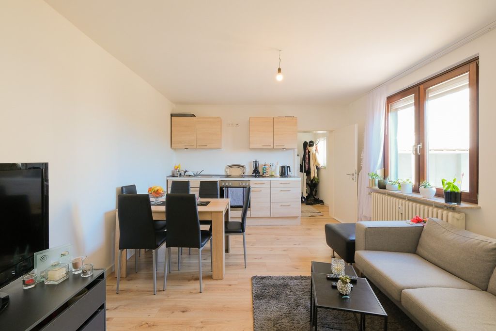 Gemütlicher Wohn- und Essbereich mit Küchenzeile, Esstisch mit schwarzen Stühlen, Sofa und großem Fenster mit Pflanzen auf der Fensterbank.