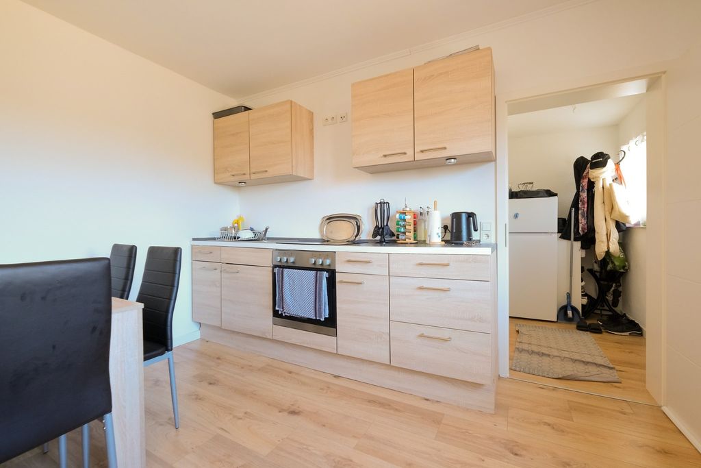 Moderne helle Küche mit Holzschränken, eingebautem Ofen und einer angrenzenden kleinen Diele mit Garderobe und Kühlschrank.