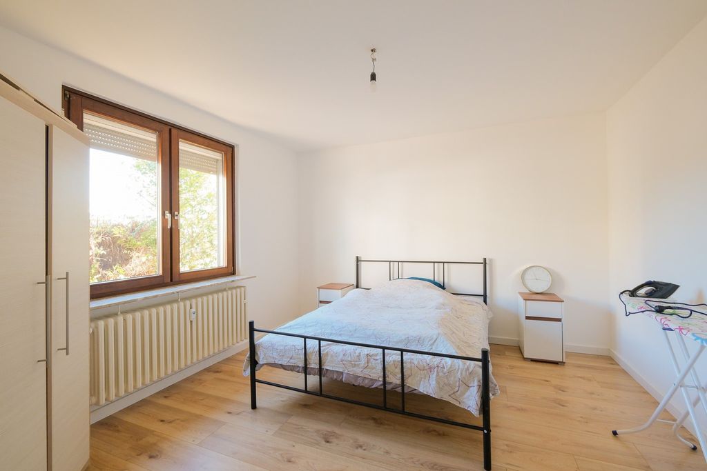 Ein schlicht eingerichtetes Schlafzimmer mit einem Doppelbett, zwei Nachttischen, einem großen Fenster und einem Bügelbrett mit Bügeleisen.