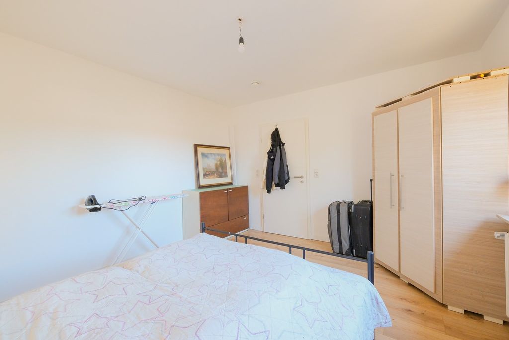 Schlafzimmer mit einem Bett mit einer weißen Bettdecke, einem Kleiderschrank, einem Kommode mit einem eingerahmten Bild, einem Bügelbrett an der Wand und zwei Koffern neben der Tür mit zwei Jacken.