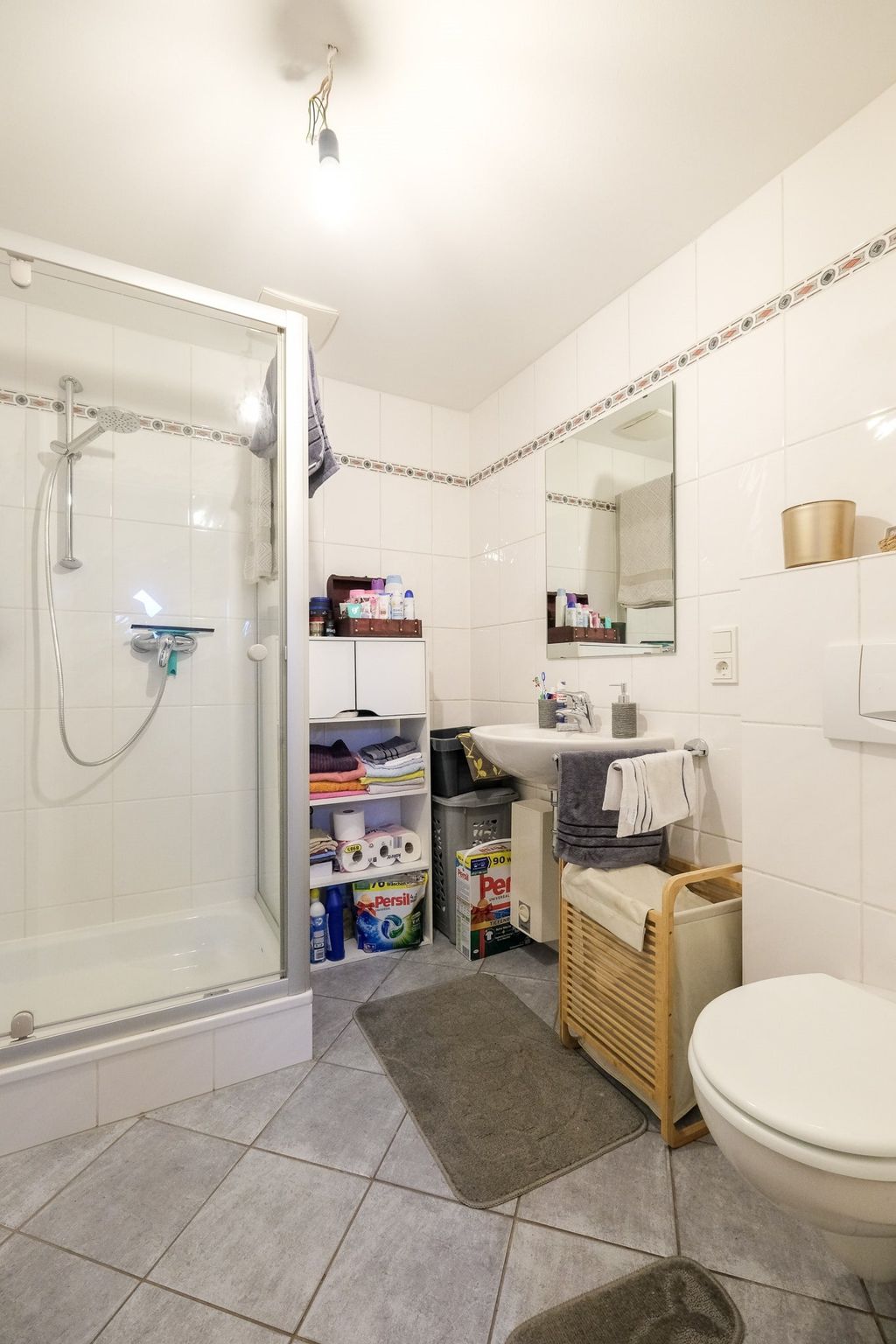 Kleines weiß gekacheltes Badezimmer mit einer Dusche, einem Waschbecken, einem WC, einem Spiegelschrank und einem Aufbewahrungsregal mit Handtüchern und Waschmitteln.