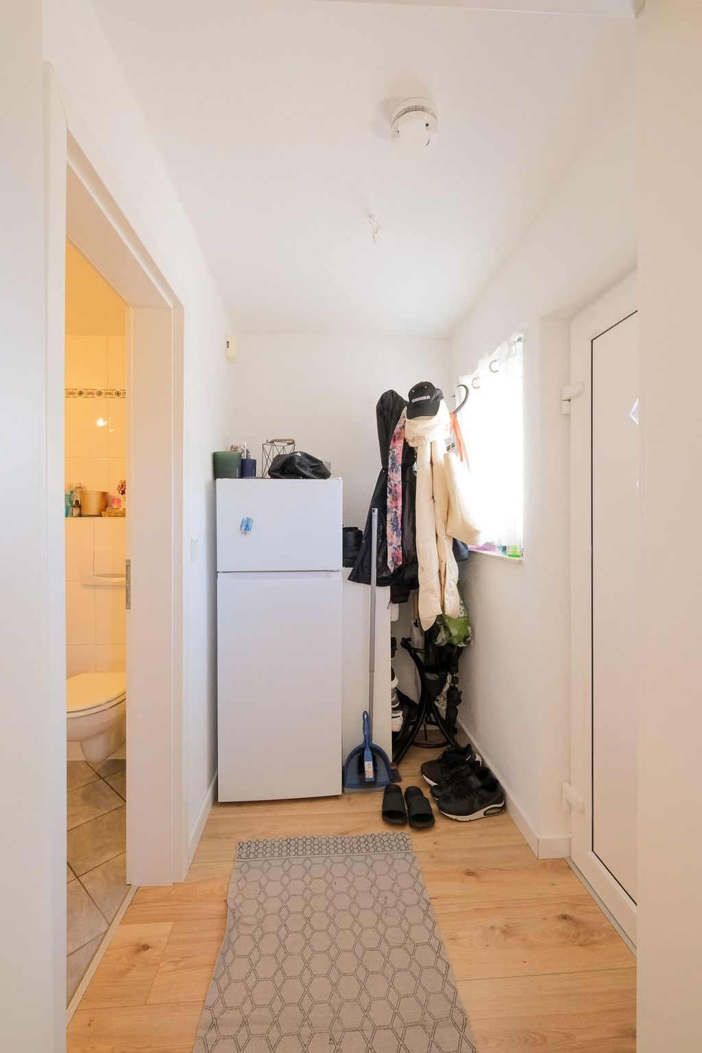 Schmaler Flur mit weißem Kühlschrank, Garderobe mit Jacken und Schuhen sowie Zugang zu einem hell erleuchteten Badezimmer.