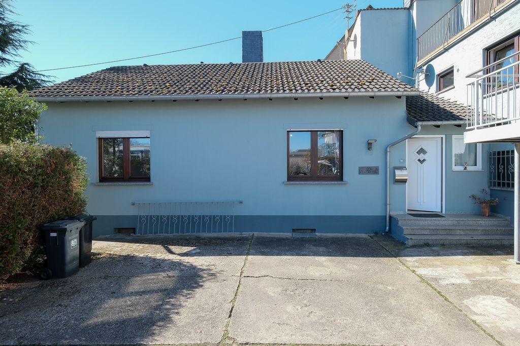 Einfamilienhaus mit hellblauer Fassade, zwei Fenstern, einer weißen Haustür rechts und Mülltonnen links auf einem betonierten Vorplatz unter blauem Himmel.