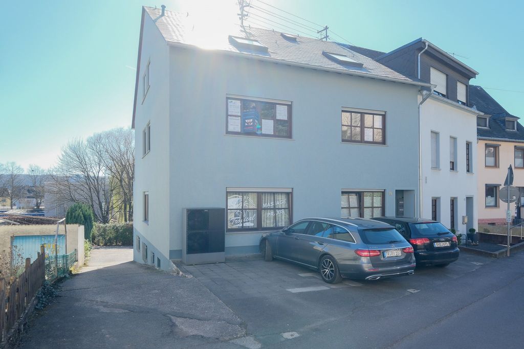Ein hellgraues Wohnhaus mit parkenden Autos auf dem Vorgartenparkplatz bei sonnigem Wetter.