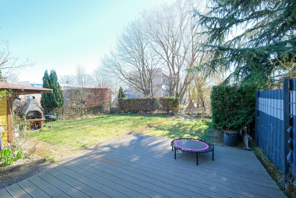 Ein sonniger Garten mit einer Holzterrasse, einem kleinen Trampolin, hohen Bäumen und einem Grillplatz in der linken Ecke.