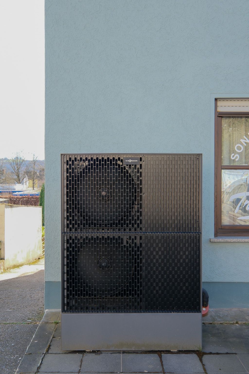 Eine schwarze Viessmann Wärmepumpe steht vor einer hellblauen Hauswand neben einem Fenster mit Vorhängen.