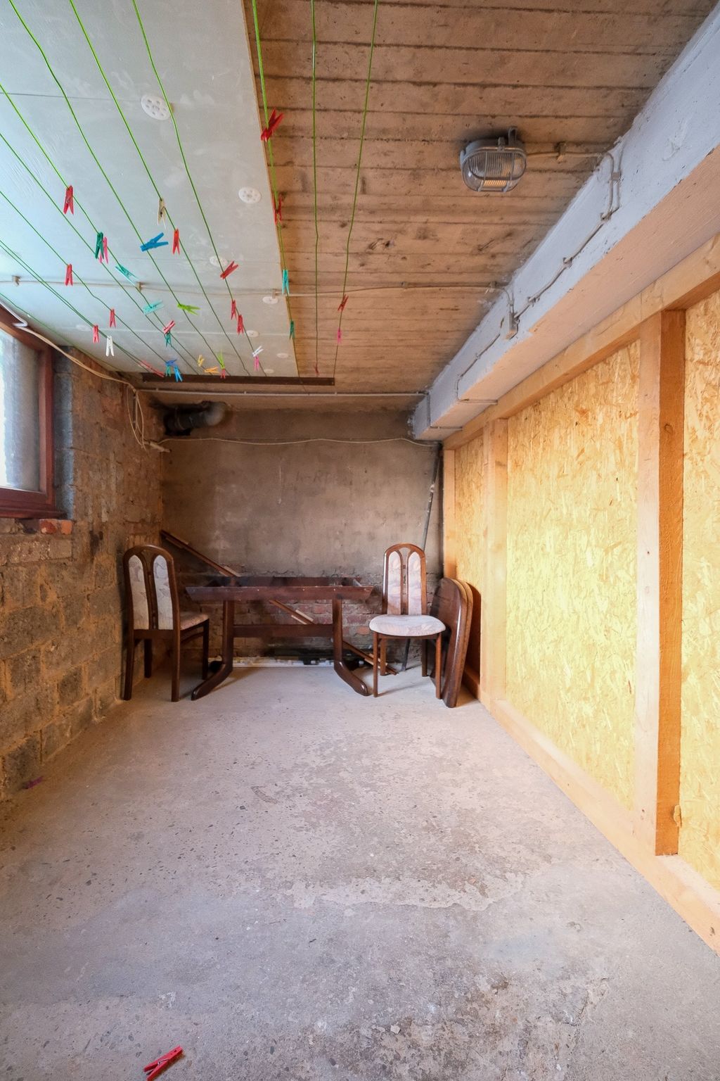 Ein spärlich eingerichteter Raum mit Betonboden, Holzstühlen und einem Tisch, mit Wäscheleinen an der Decke und einer halboffenen Fensterseite.