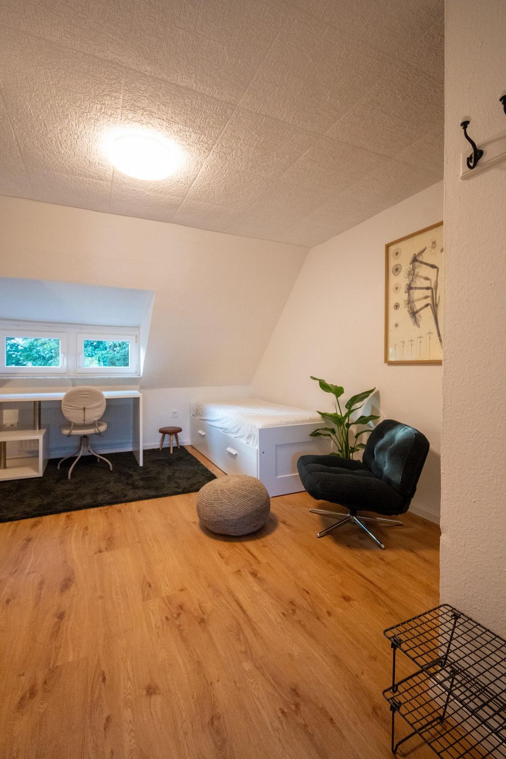 Ein minimalistisches Zimmer mit hellen Wänden, einem weißen Bett, einem Schreibtisch mit einem Stuhl vor einem Fenster, einem schwarzen Sessel neben einer grünen Pflanze und einem grauen Pouf auf einem Holzfußboden.