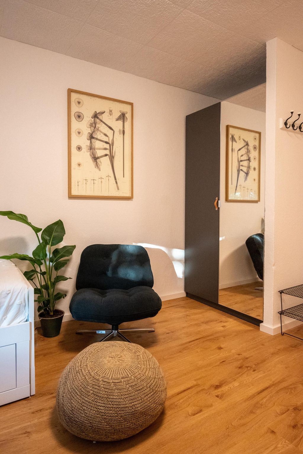 Ein gemütlicher Raum mit einem dunkelblauen Sessel, einem runden gestrickten Ottoman vor einem Kleiderschrank mit Spiegeltür, einer Topfpflanze neben einem weißen Bett und einer gerahmten botanischen Wandzeichnung.
