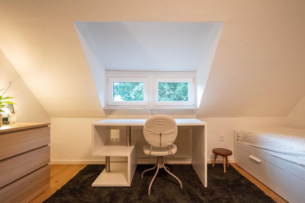 Helles, minimalistisches Zimmer mit einem weißen Schreibtisch vor einem Dachfenster, einem grauen Bürostuhl, einem kleinen Hocker und einem Bett mit weißen Schubladen unter der Schräge.