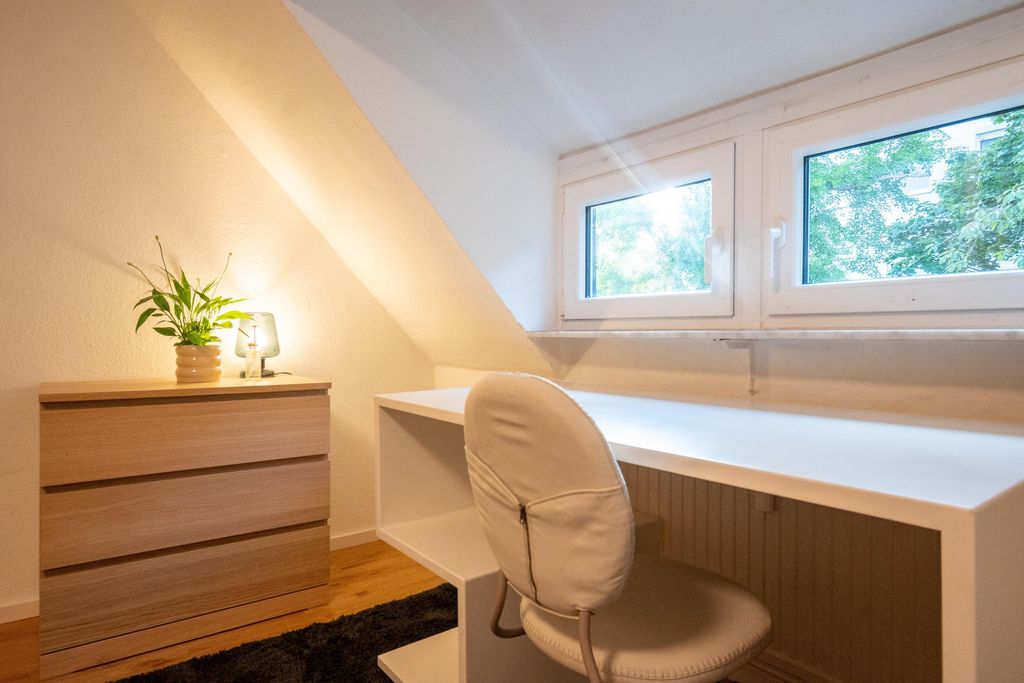 Helles Zimmer mit weißem Schreibtisch, grauem Stuhl und einem kleinen Holzdrehschrank mit Pflanze und Lampe unter einer Dachschräge.