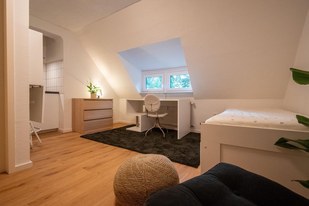 Ein schlicht eingerichtetes Zimmer mit schräger Decke, einem weißen Schreibtisch vor einem kleinen Fenster, einem weißen Stuhl, einem Bett rechts und einer Holzkommode mit einer Pflanze.