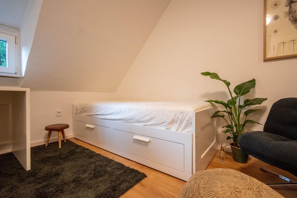 Ein schlichtes Schlafzimmer unter einem schrägen Dach mit einem weißen Bett, schwarzem Teppich, kleinem Hocker, Topfpflanze und einem dunklen Sessel.