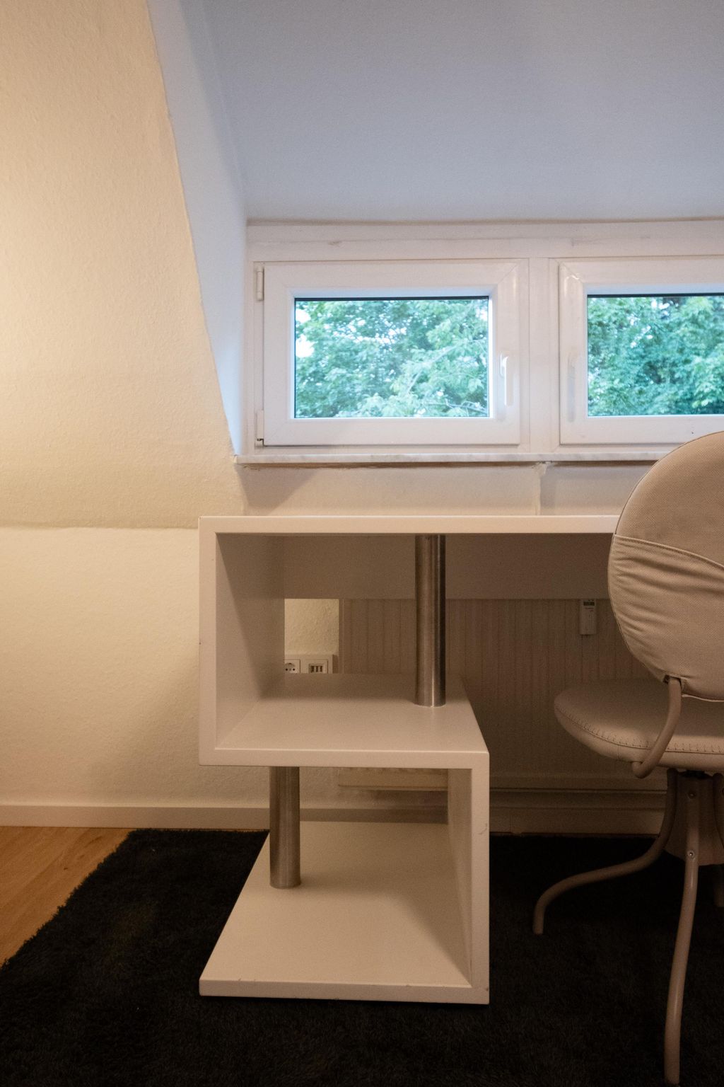 Weißes, modernes, geometrisch geformtes Beistelltischregal aus Holz mit Metallstützen vor einem kleinen Fenster mit grünen Bäumen draußen.