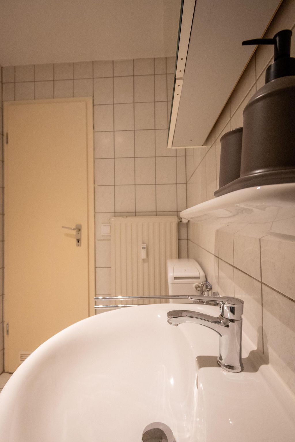 Ein Badezimmer mit weißem Waschbecken und silbernem Wasserhahn, Wand mit quadratischen Fliesen, beige Türe, Heizkörper und Seifenspender auf der rechten Seite.