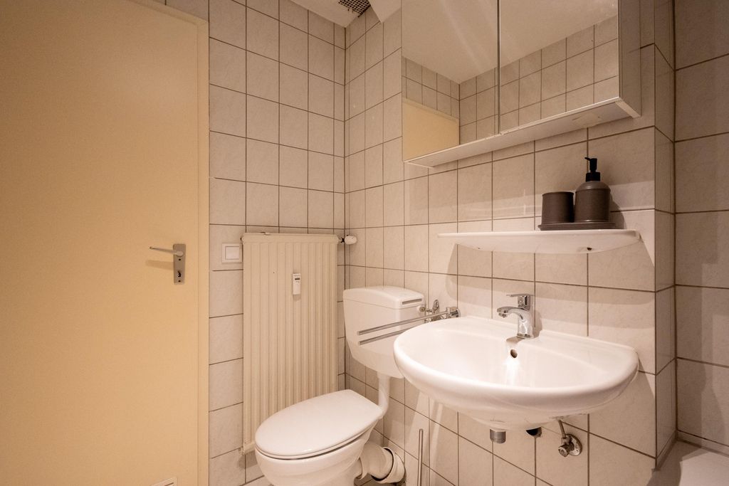 Kleines Badezimmer mit weißem WC und Waschbecken, beige Fliesen an den Wänden. An der Wand hängt ein Spiegel mit einem Regal, auf dem zwei Seifenspender stehen.