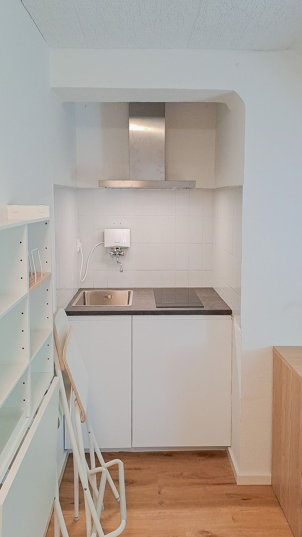 Kleine Küchenzeile mit weißer Fliesenspiegelwand, Spüle, Herd und Edelstahl-Dunstabzugshaube in einer Nische, vor der sich zwei zusammengeklappte Stühle befinden.