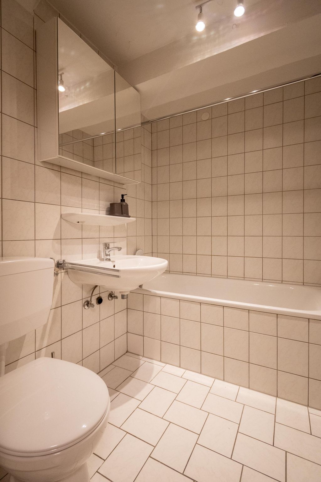 Kleines Badezimmer mit weißen Fliesen, Toilette, Wandwaschbecken mit Armatur, halbrundem Spiegeloberschrank, Seifenspender und Badewanne.