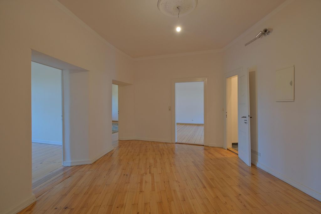 Leerer Raum mit Holzfußboden und weißen Wänden, vier offene Türöffnungen zu angrenzenden Zimmern, eine Glühbirne an der Decke.