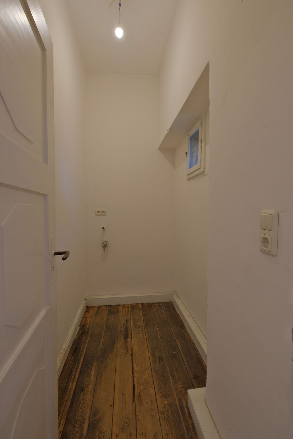 Kleiner, leerer Raum mit weißen Wänden, einem kleinen Fenster und einem hängenden Licht in der Decke, mit einem Holzboden und teilweise sichtbarer geöffneter Tür.