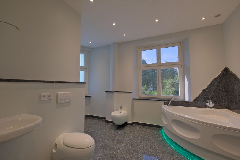 Ein modernes Badezimmer mit grauem Fliesenboden, weißen Wänden und einer Eckbadewanne mit grüner LED-Beleuchtung, Waschbecken, Toilette und Bidet, vor einem großen Fenster mit Blick ins Grüne.
