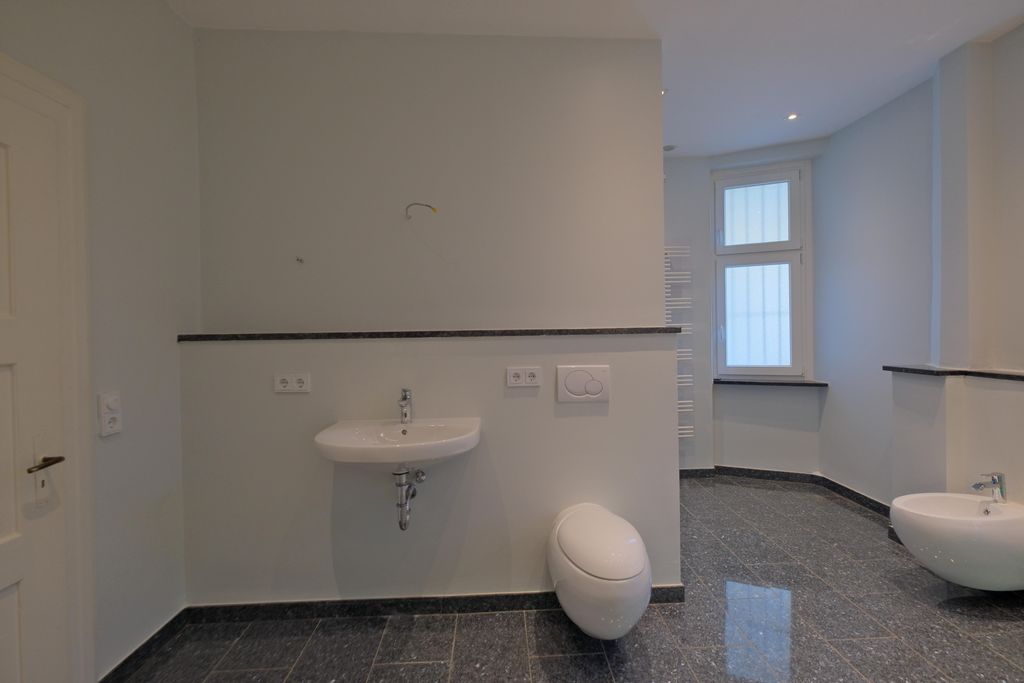 Ein modernes Badezimmer mit weißen Wänden, einer hängenden Toilette, einem Wandwaschbecken und dunklem Fliesenboden. Ein Fenster und ein Heizkörper sind im Hintergrund sichtbar.