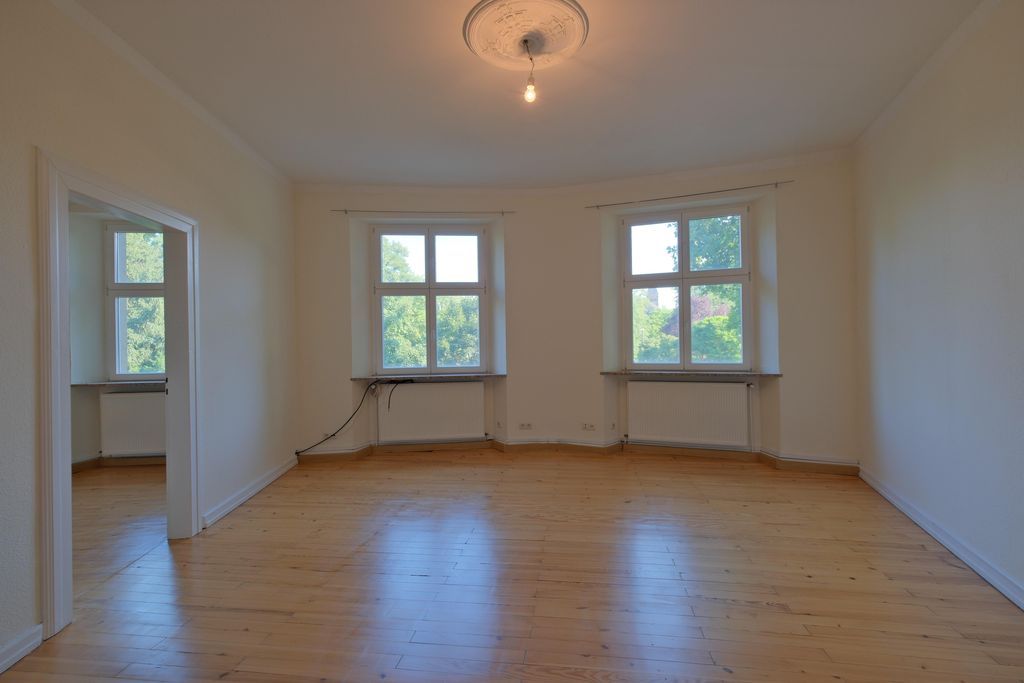 Leerer Raum mit hellen Holzfußböden, zwei großen Fenstern mit Blick auf Bäume und eine angrenzende Tür zu einem weiteren Raum mit Fenstern.