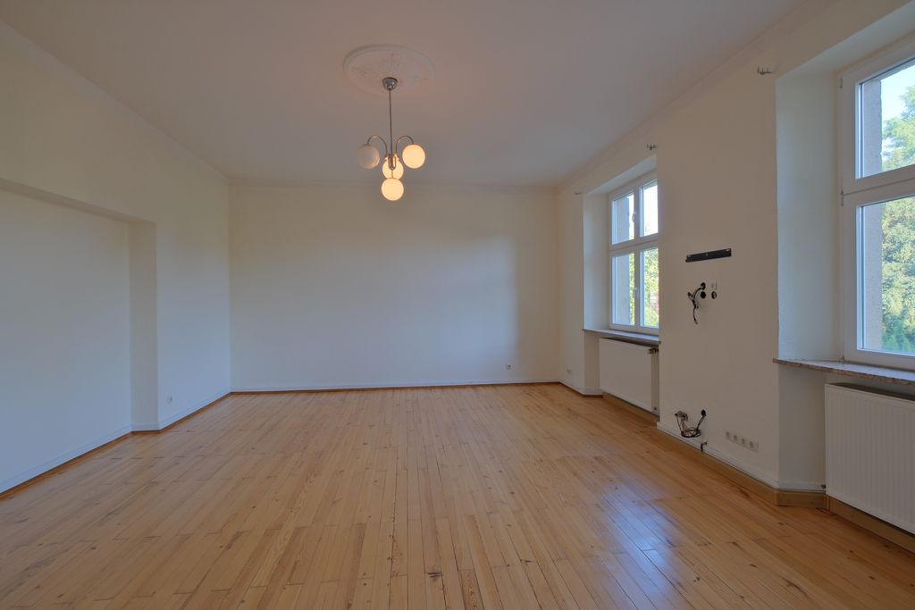 Leerer heller Raum mit Holzboden, großen Fenstern auf der rechten Seite und einer modernen Deckenleuchte.
