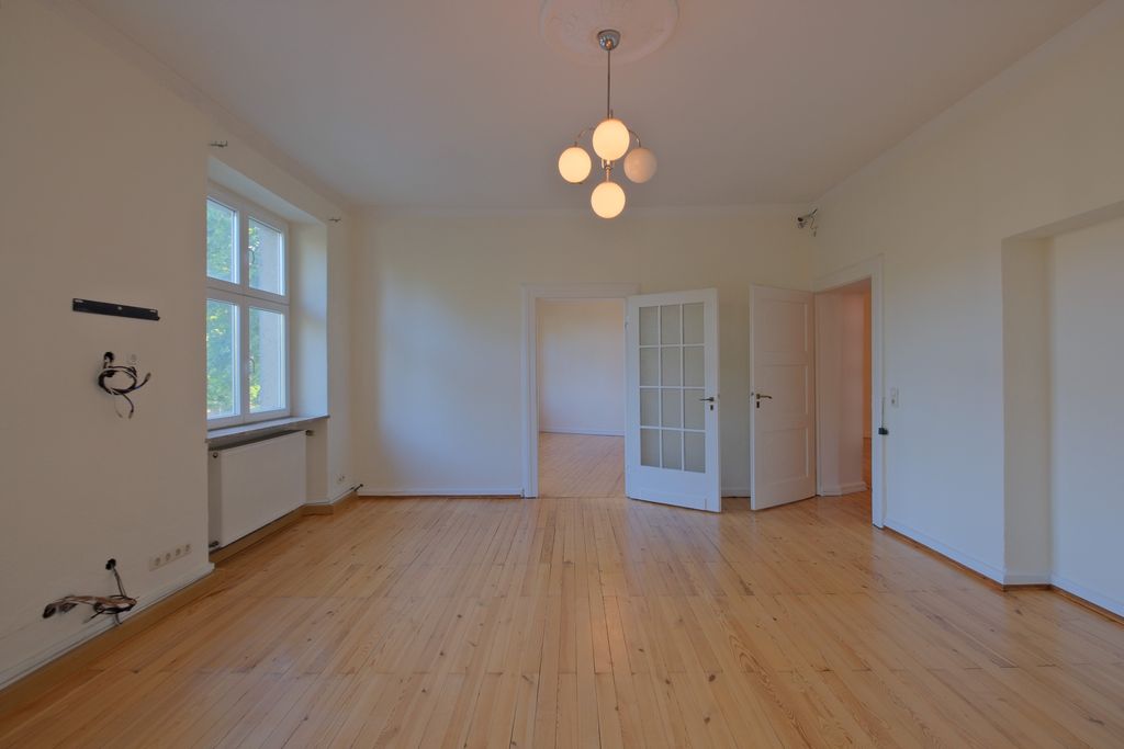 Leerer Raum mit hellem Holzboden, weißen Wänden und einer modernen Deckenleuchte mit fünf runden Lampen, sichtbare Türen und Fenster mit Blick nach draußen.