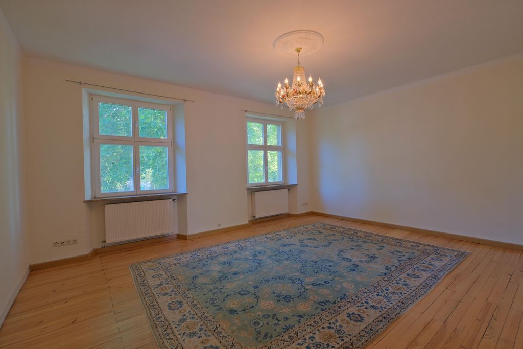Leerer Raum mit hellen Wänden, zwei Fenstern, die Tageslicht und grüne Bäume draußen zeigen, einem großen dekorativen Teppich auf dem Holzfußboden und einem Kronleuchter an der Decke.
