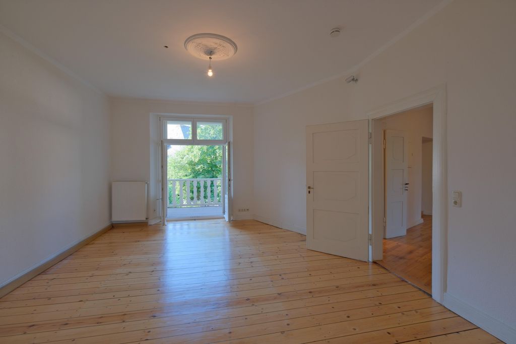 Leerer Raum mit Holzboden, offenem Balkontürfenster mit Blick auf grüne Bäume und weißen Wänden.