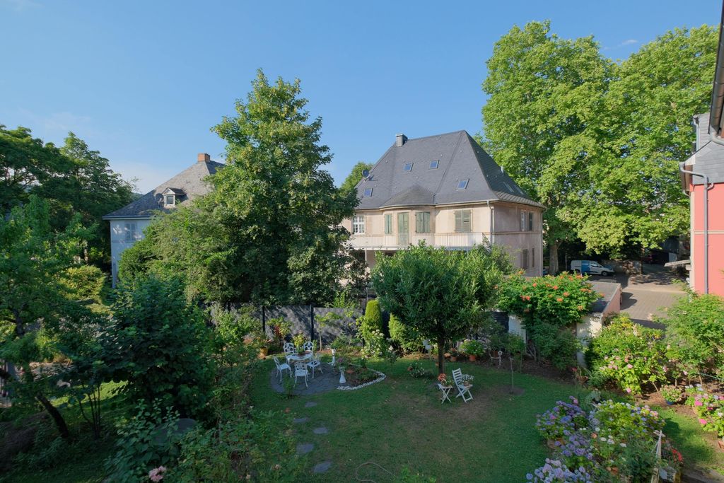 Blick auf einen gepflegten Garten mit verschlungenen Wegen, weißen Gartenmöbeln und blühenden Pflanzen vor einem großen, historischen Haus mit einem steilen, dunklen Dach und grünen Fensterläden unter blauem Himmel.