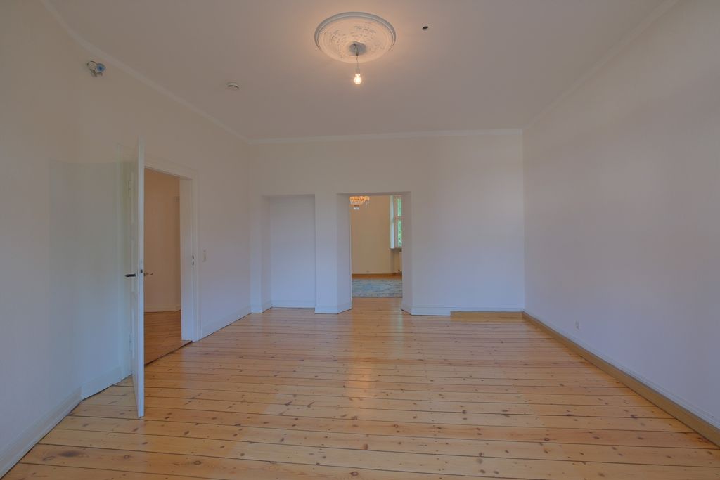 Leerer Raum mit weißen Wänden und Holzboden, ausgestattet mit einer einzelnen Deckenlampe und offenen Türen, die in einen weiteren Raum führen.