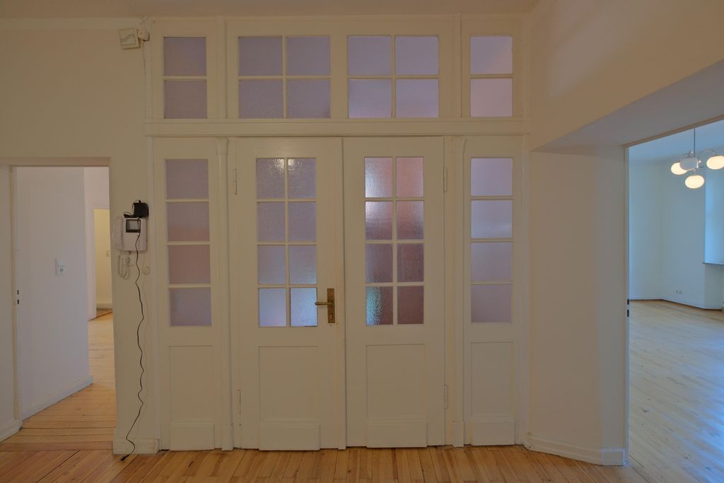 Blick auf eine weiße Doppeltür mit verglasten Fensterabschnitten in einem hellen Raum mit Holzfußboden. Links und rechts der Tür sind offene Durchgänge zu weiteren Räumen sichtbar.