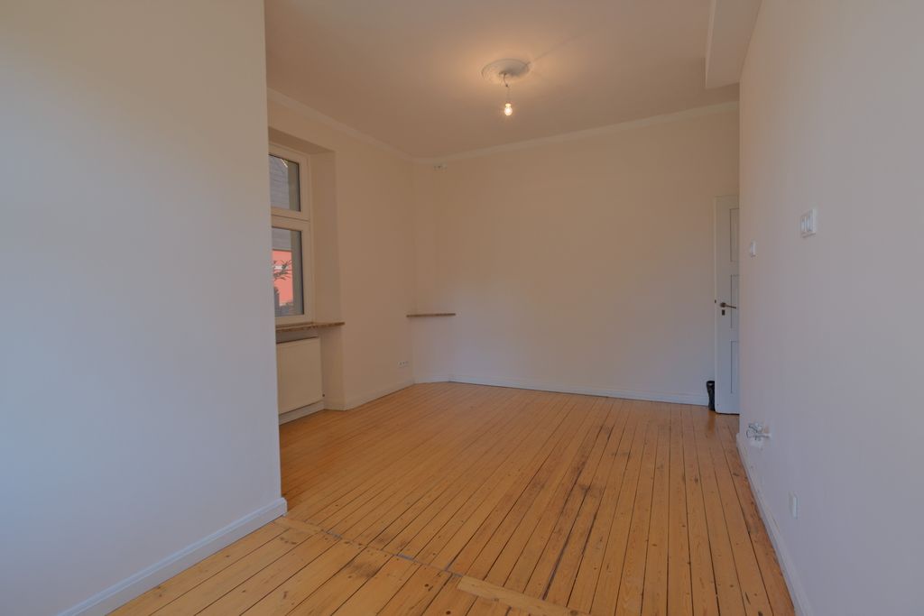 Leerer Raum mit weißen Wänden, großen Fenstern und einem Boden aus hellem Holz. Eine nackte Glühbirne hängt von der Decke.