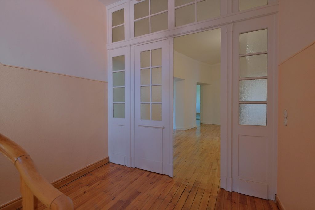 Offene weiße Glastüren führen von einem Flur mit Holzgeländer in einen Raum mit Holzboden und weißen Wänden.