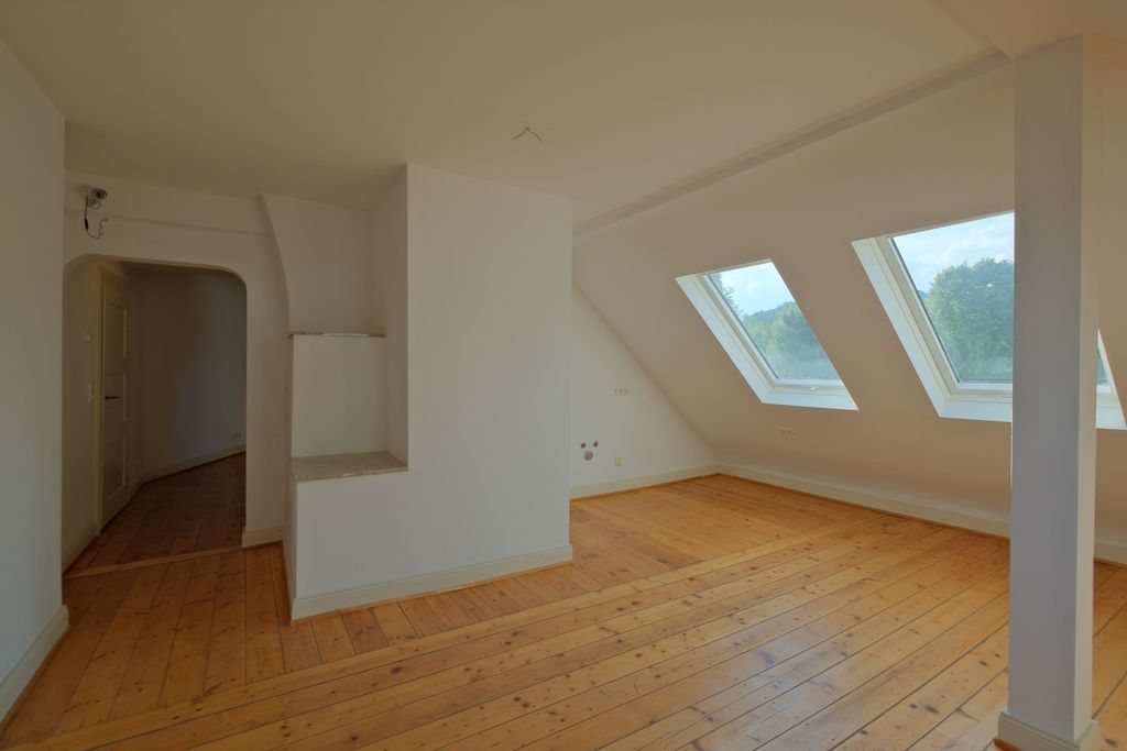 Helles, leeres Dachzimmer mit Holzboden, drei großen Dachfenstern und sichtbarem Durchgang zu einem Flur mit weißen Wänden.